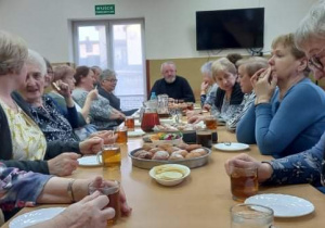 Seniorzy z Klubu "Senior+" siedzą przy stole i degustują upieczone przez siebie pączki.